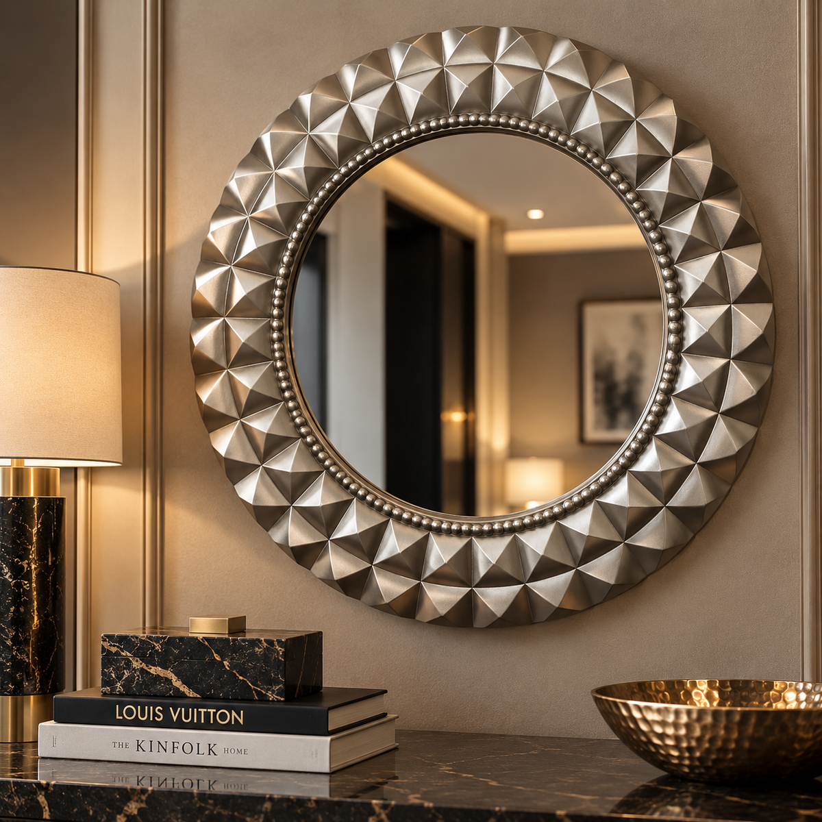 A large round wall mirror with a textured, faceted metallic frame in a champagne-gold finish, mounted on a beige wall above a dark marble console table with books and a lamp.