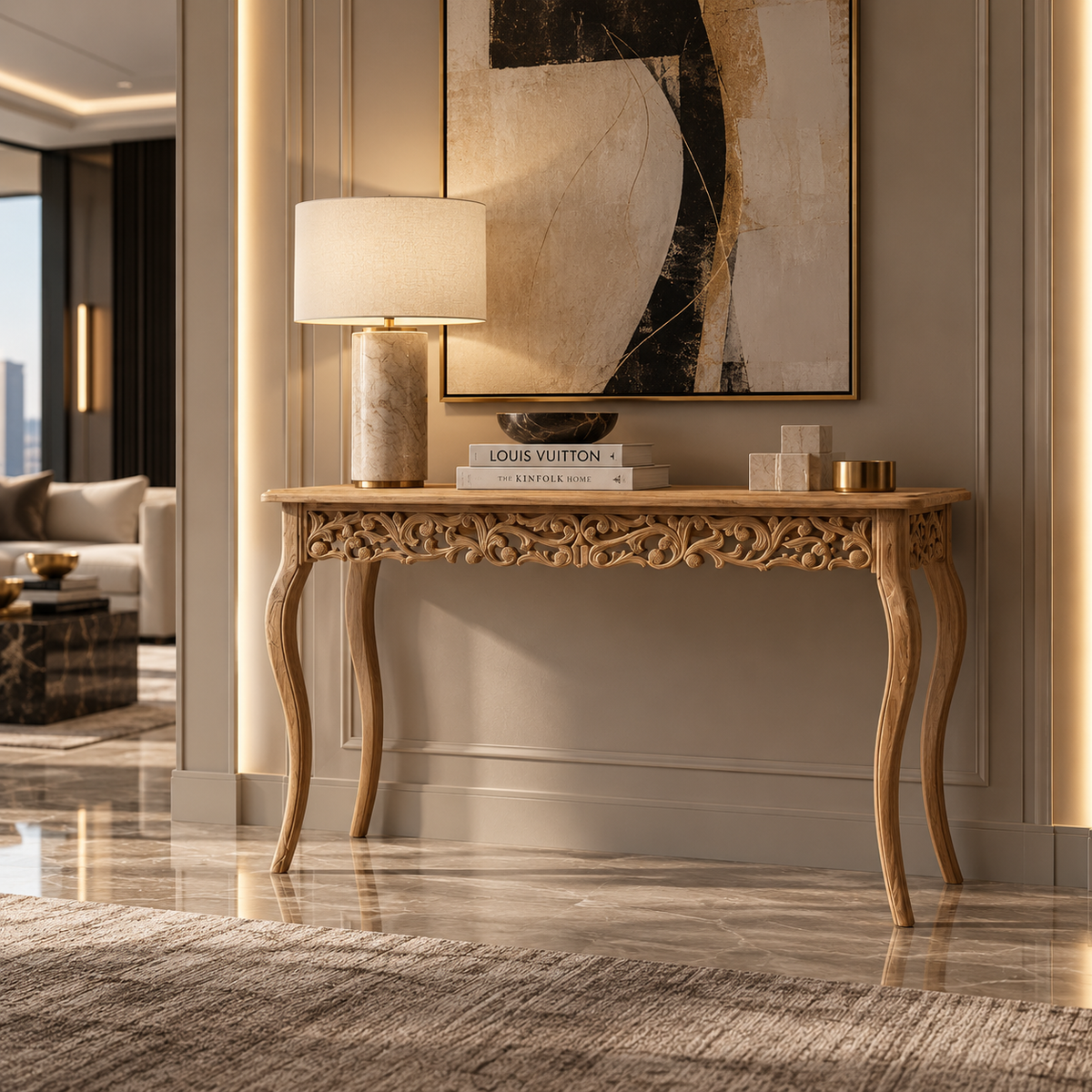 An ornate carved wooden console table in a warm light oak finish, styled in a luxury modern living room setting with a table lamp, books, and decorative objects on top.