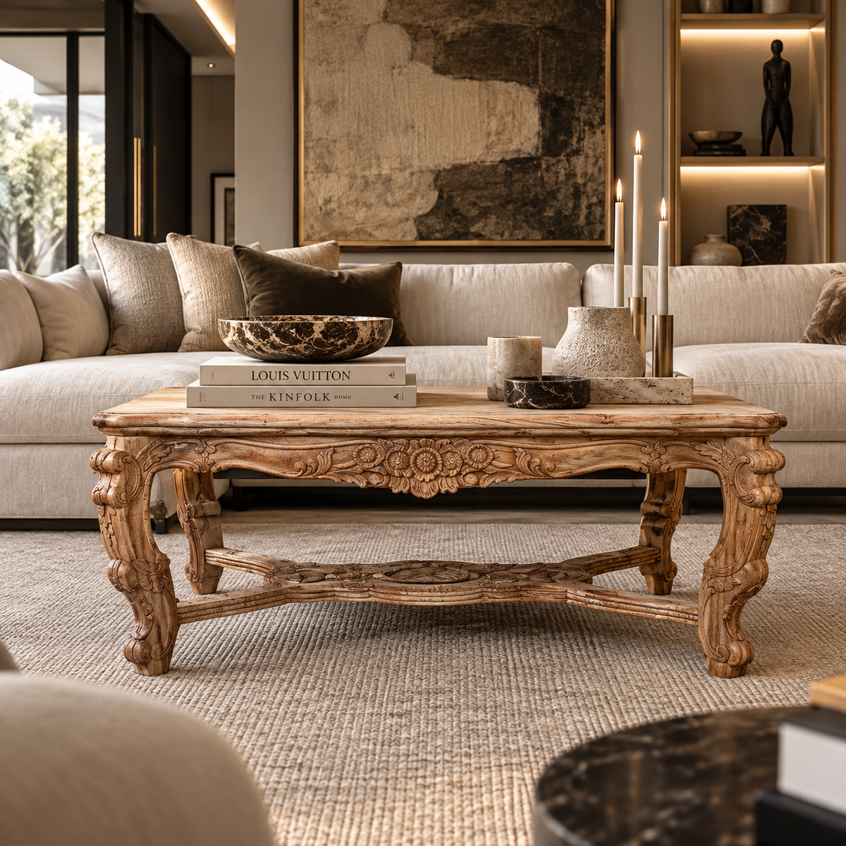 An ornate carved wooden coffee table in a styled living room setting, decorated with books, a bowl, and candles, positioned in front of a cream-colored sofa.