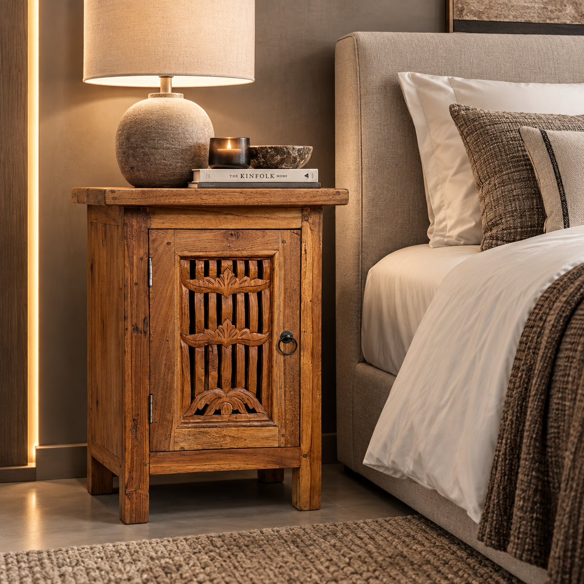 A rustic wooden nightstand with carved details and a single cabinet door, styled next to a bed in a warm, modern bedroom setting.