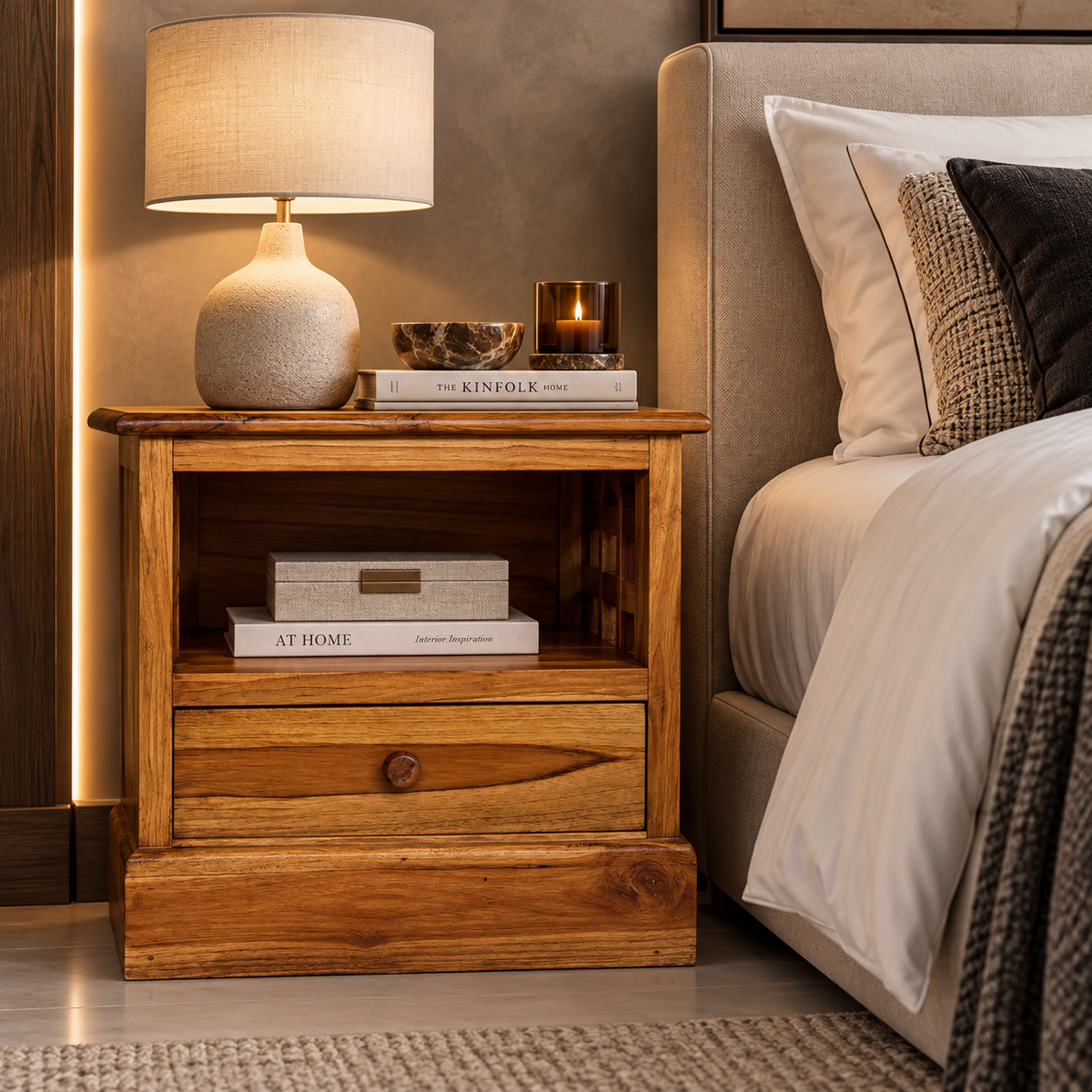 A rustic wooden nightstand with a single drawer and open shelf, styled in a bedroom next to a bed with white linens and a lit table lamp on top.