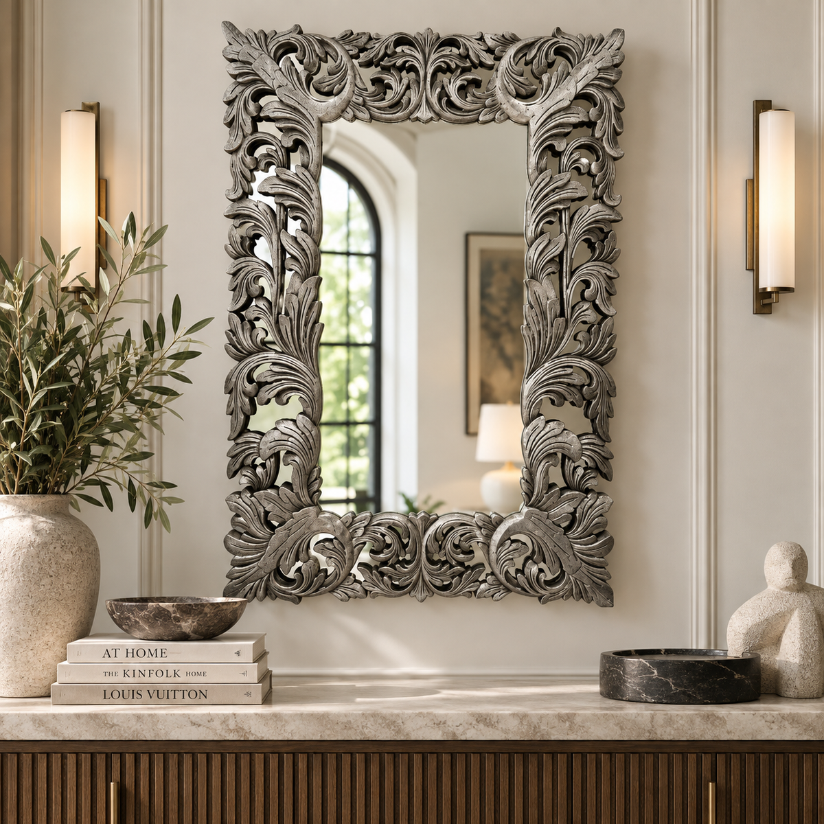 An ornate rectangular wall mirror with a highly detailed, carved silver-gray floral and scrollwork frame, displayed in a styled interior setting above a console table.