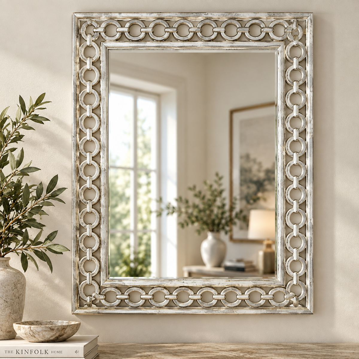 A large rectangular wall mirror with an ornate distressed silver frame featuring interlocking circular motifs, styled on a wooden table next to a vase with greenery.