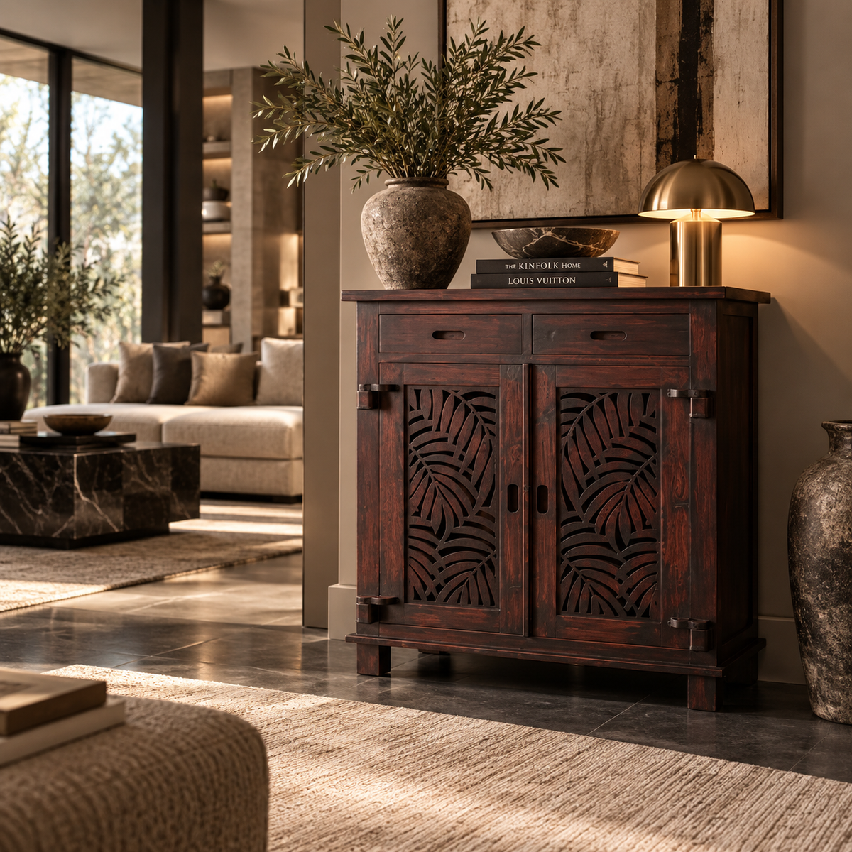 A dark reddish-brown wooden sideboard cabinet with carved leaf-pattern doors, styled in a modern living room with a vase, books, and a lamp on top.