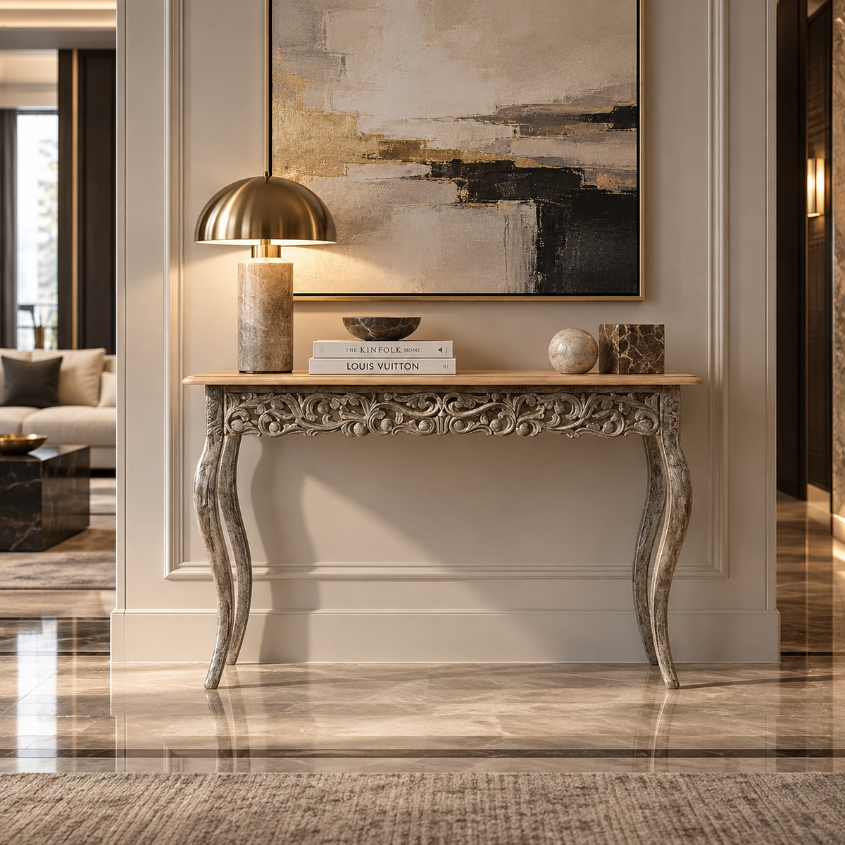 An ornate carved console table in a warm beige and gray finish stands against a cream wall in a luxury interior. A brass dome lamp, stacked books including one labeled Louis Vuitton, a small bowl, and decorative vases sit on the tabletop.