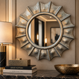 A decorative sunburst-style wall mirror with an antiqued metallic frame hangs on a beige wall above a dark marble console. A lamp, stacked books including Louis Vuitton, and a hammered gold bowl are visible in the scene.