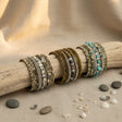 Collection of handcrafted multi-layer wrap bracelets featuring turquoise, ivory, bronze, silver, and natural stone beads, displayed on a rustic driftwood branch with decorative shells and stones on a neutral linen surface.