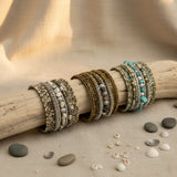 Collection of handcrafted multi-layer wrap bracelets featuring turquoise, ivory, bronze, silver, and natural stone beads, displayed on a rustic driftwood branch with decorative shells and stones on a neutral linen surface.