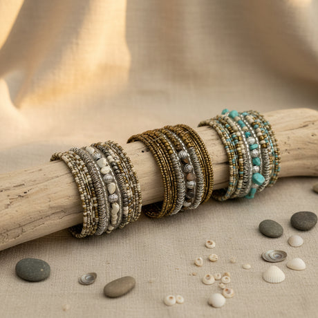 Collection of handcrafted multi-layer wrap bracelets featuring turquoise, ivory, bronze, silver, and natural stone beads, displayed on a rustic driftwood branch with decorative shells and stones on a neutral linen surface.