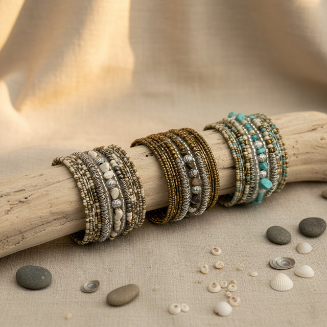 Collection of handcrafted multi-layer wrap bracelets featuring turquoise, ivory, bronze, silver, and natural stone beads, displayed on a rustic driftwood branch with decorative shells and stones on a neutral linen surface.