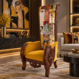An ornate high-back accent chair with carved dark wood arms and legs, a mustard yellow velvet seat, and a tall backrest covered in a patchwork collage print, shown in a styled living room setting.
