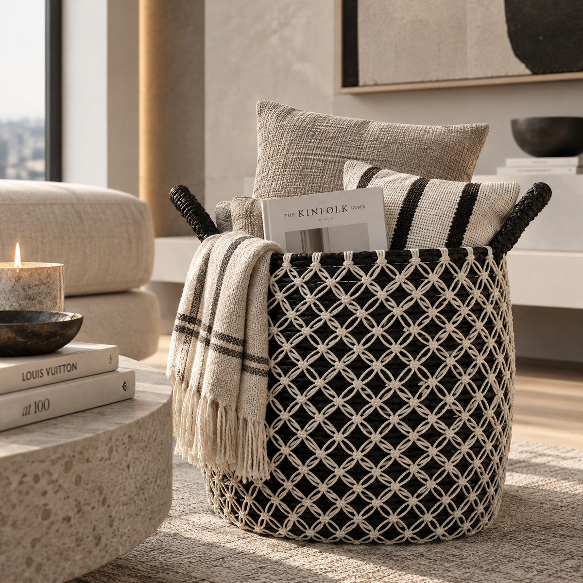 A black and cream woven basket with black rope handles, styled in a modern living room setting with a cream throw blanket, pillows, and books.