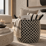 A black and cream woven basket with black rope handles, styled in a modern living room setting with a cream throw blanket, pillows, and books.