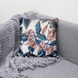 Decorative scatter cushion featuring soft blush florals and deep blue botanical leaves on a neutral background, styled on a grey sofa with a textured throw.
