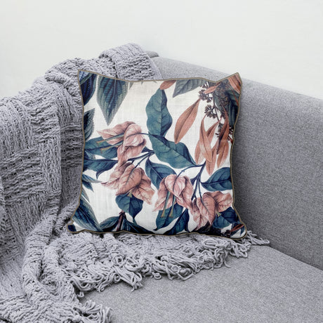 Decorative scatter cushion featuring soft blush florals and deep blue botanical leaves on a neutral background, styled on a grey sofa with a textured throw.