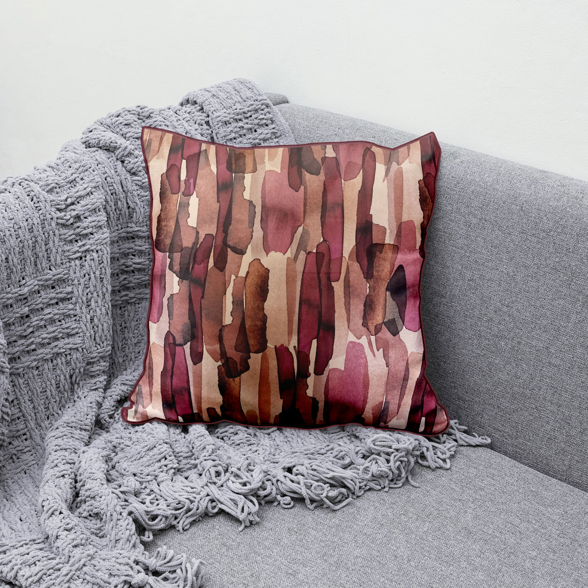 Decorative scatter cushion featuring a watercolour-inspired abstract pattern in blush pink, burgundy, rust, brown, and soft neutral tones, styled on a grey sofa.