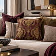A decorative square throw pillow with an ornate gold and burgundy damask pattern, styled on a beige textured sofa alongside other accent pillows in a modern living room setting.