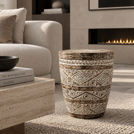 A round ceramic stool with a rustic carved geometric pattern in beige and brown sits on a woven rug in a modern living room setting.
