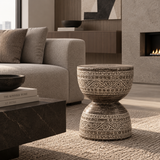 A round two-tiered pedestal stool with a textured beige and brown bohemian-style geometric pattern, styled in a modern living room next to a gray sofa and a dark marble coffee table.