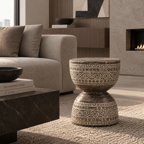 A round two-tiered pedestal stool with a textured beige and brown bohemian-style geometric pattern, styled in a modern living room next to a gray sofa and a dark marble coffee table.
