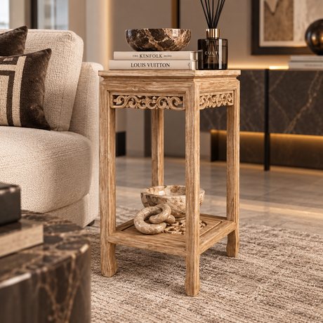 A small carved wooden end table in a warm light oak finish, styled in a modern living room next to a beige sofa. The table features ornate scrollwork carvings on the upper frame and a lower shelf, holding a stack of books, a decorative bowl, and a reed diffuser.