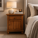 A warm medium-brown wooden nightstand with a single drawer and a cabinet door, styled in a bedroom next to a bed. On top of the nightstand sits a lit table lamp, a stack of books, and a small candle.