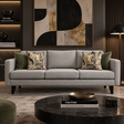A modern three-seat sofa in light gray fabric with black legs, styled in a contemporary living room. The sofa is decorated with two olive green throw pillows and two patterned floral pillows, set against a dark wall with a large abstract art piece and a round black marble coffee table in the foreground.