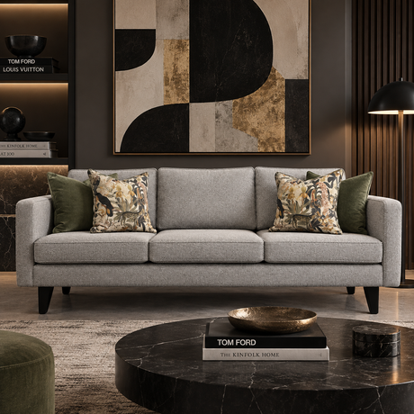 A modern three-seat sofa in light gray fabric with black legs, styled in a contemporary living room. The sofa is decorated with two olive green throw pillows and two patterned floral pillows, set against a dark wall with a large abstract art piece and a round black marble coffee table in the foreground.