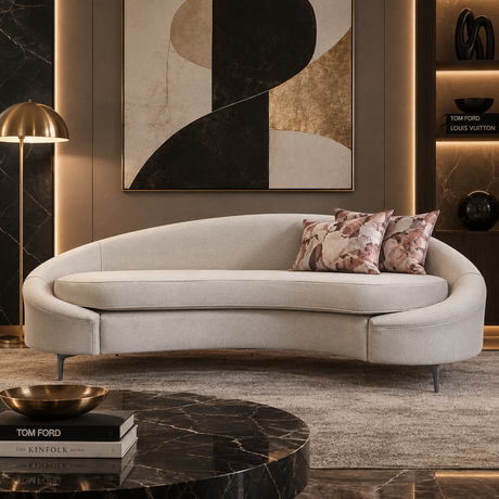 A curved upholstered sofa in a light warm gray or beige fabric, styled in a luxury interior with two floral throw pillows, a gold floor lamp, a marble coffee table, and a large abstract wall art piece.