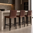 Three bar stools with dark brown woven seats and tall backrests, black wooden legs, and rectangular footrests beside a kitchen island.