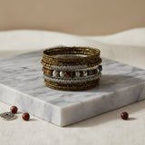 Handcrafted multi-layer wrap bracelet featuring bronze seed beads, silver textured beads, and tiger's eye stone accents, displayed on a white marble block against a warm neutral background.