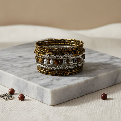 Handcrafted multi-layer wrap bracelet featuring bronze seed beads, silver textured beads, and tiger's eye stone accents, displayed on a white marble block against a warm neutral background.