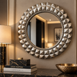 A large round wall mirror with a textured, faceted metallic frame in a champagne-gold finish, mounted on a beige wall above a dark marble console table with books and a lamp.