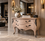 A light natural wood French console table with carved black metal accents and curved legs, styled in a luxurious interior setting with a marble vase of white blossoms and decorative books on top.