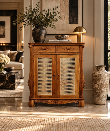 An ornate mid-century wooden cabinet with woven cane doors, styled in a warm, modern living room with a brass table lamp, ceramic vases, and a marble side table.