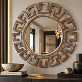 Round decorative wall mirror with a distressed brown Greek key frame, mounted on a beige wall above a console table with decorative bowls and vases.