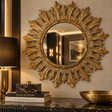 A large round sunburst wall mirror with an ornate gold leaf design, mounted on a beige wall above a dark marble console table with a lamp, books, and a bowl.