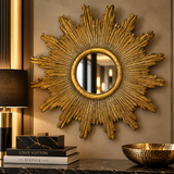 A large gold sunburst wall mirror with a textured, metallic starburst frame is mounted on a beige wall above a dark marble console. The scene is styled with a Louis Vuitton book, a black lamp, and a hammered gold bowl.
