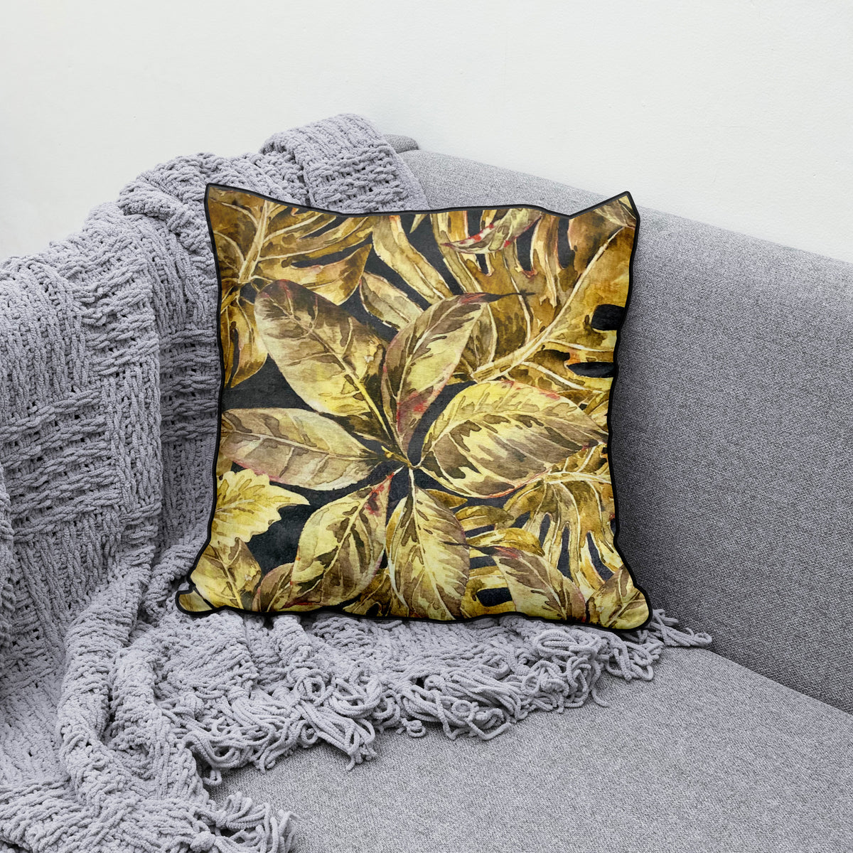 Decorative scatter cushion featuring a rich tropical leaf pattern in gold, mustard, olive, and charcoal tones with dark piping, styled on a textured grey sofa.