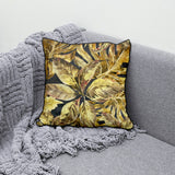Decorative scatter cushion featuring a rich tropical leaf pattern in gold, mustard, olive, and charcoal tones with dark piping, styled on a textured grey sofa.