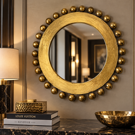 Round decorative wall mirror with a textured gold frame and a border of metallic gold beads, mounted on a beige wall above a marble console table with books and a lamp.