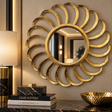 A large decorative wall mirror with a sunburst gold frame hangs on a beige wall above a dark marble console. A lit lamp, stacked books including Louis Vuitton, and a hammered gold bowl are visible in the styled interior setting.