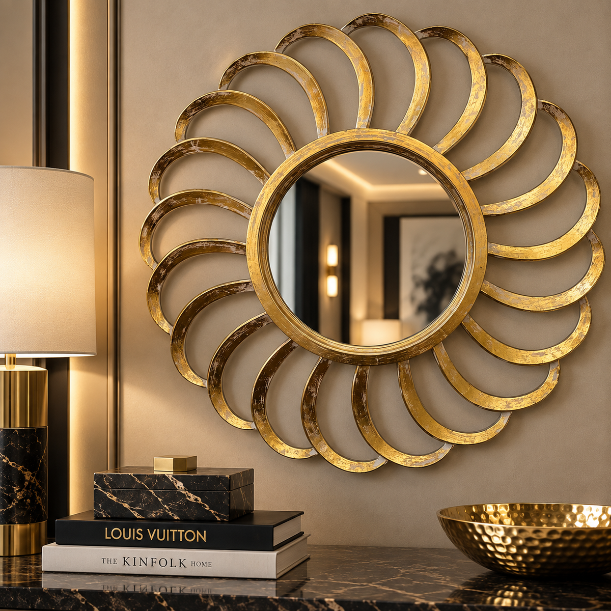 A large decorative wall mirror with a sunburst gold frame hangs on a beige wall above a dark marble console. A lit lamp, stacked books including Louis Vuitton, and a hammered gold bowl are visible in the styled interior setting.