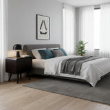 Grey upholstered bed with white bedding, teal and grey cushions, and a grey throw in a bright bedroom with a dark bedside table.