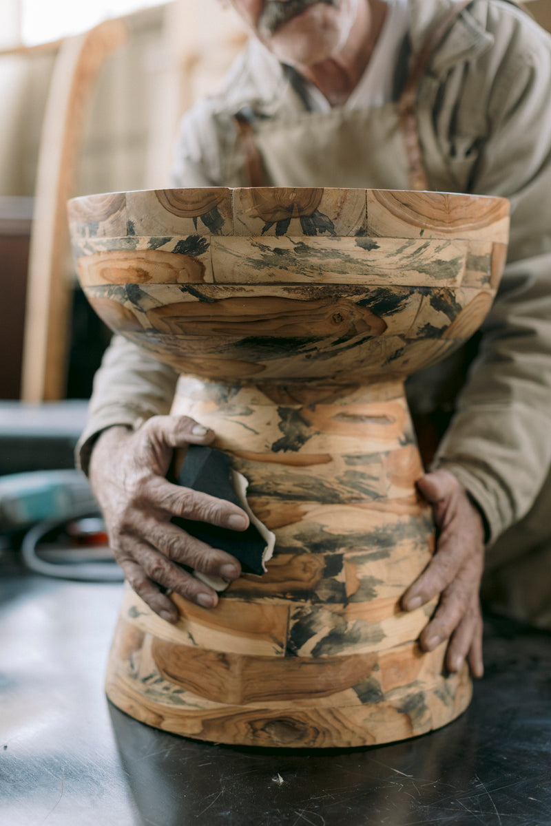 Handcrafted solid teak pedestal bowl featuring a natural wood grain with a sculptural silhouette. Shown being hand-finished by a skilled artisan, this decorative wooden bowl celebrates expert craftsmanship and timeless organic design for luxury interiors.