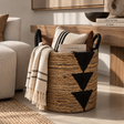 A woven natural-fiber storage basket with black handles and large black geometric triangle motifs, styled in a warm living room setting with a cream throw blanket, pillows, and books.