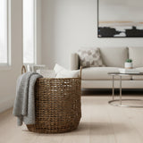 Handwoven natural fiber storage basket with integrated handles, styled with a knitted throw, decorative cushions, and books in a modern living room, offering stylish storage for blankets and home essentials.