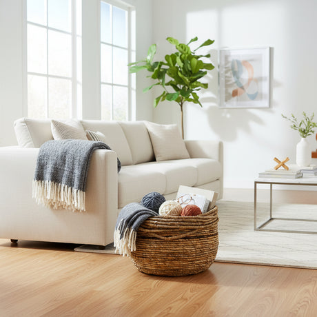 Handwoven natural storage basket with integrated carry handles, styled with blankets, yarn, and magazines beside a contemporary sofa, offering a stylish and practical storage solution while adding warm organic texture to modern, coastal, and rustic interiors.