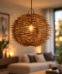 A spherical pendant light made of tightly woven natural branches or wicker, glowing with a warm light bulb inside, hanging in a softly blurred modern living room with a sofa and window in the background.