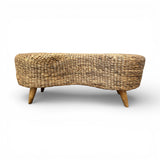 Handwoven natural rattan bench with a sculptural organic silhouette and solid wooden legs, offering a warm textured finish and stylish seating for entryways, bedrooms, living rooms, or covered outdoor spaces.